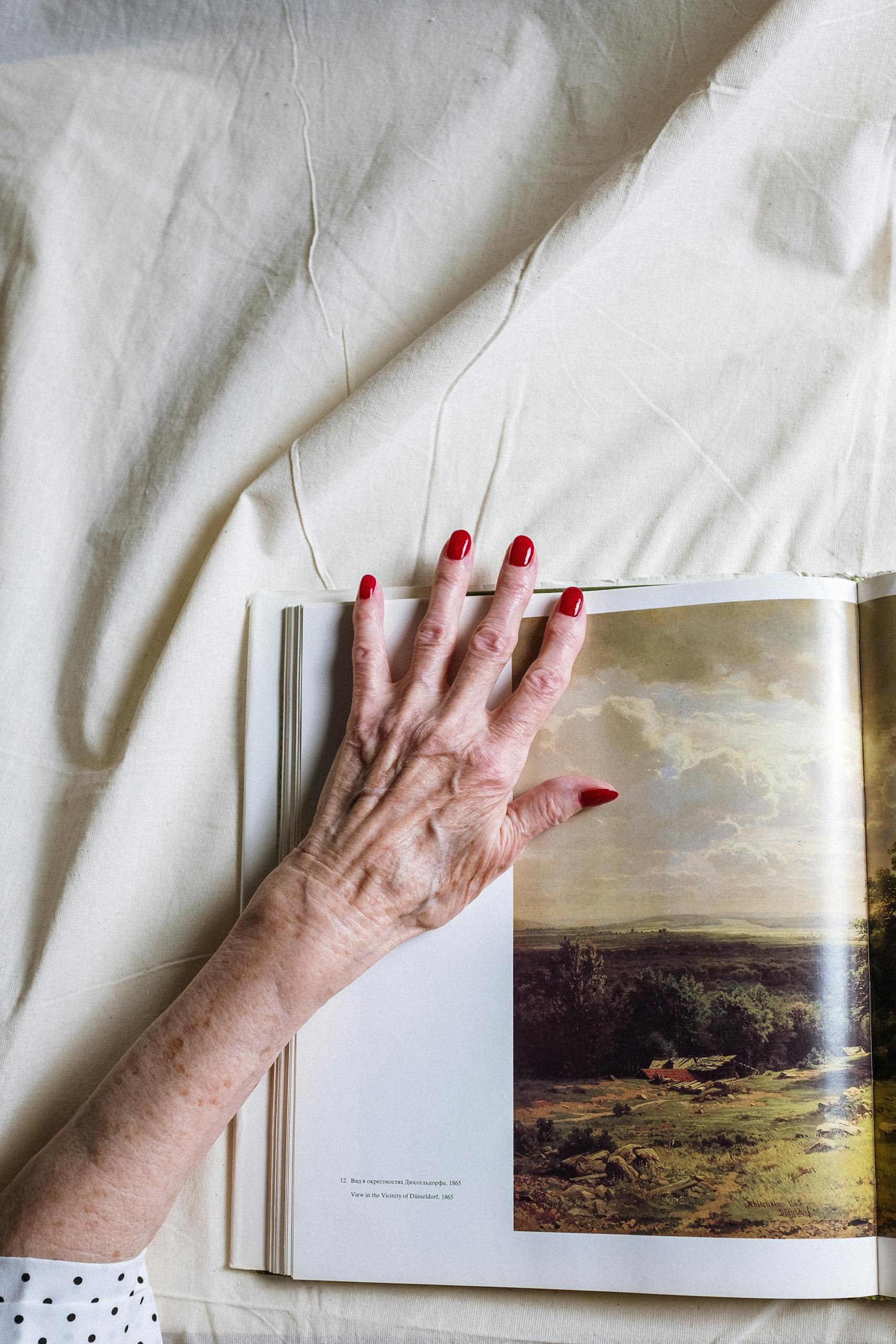 An elderly hand with red nails rests on an open book, showcasing artwork.